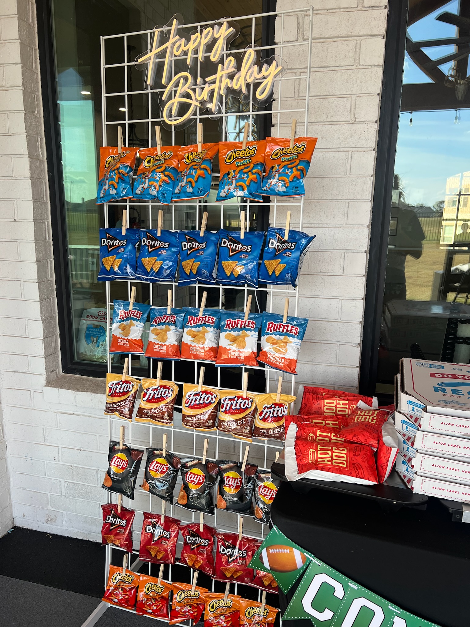 football birthday bash set up, white standing grid panel wall holding snack-sized chip bags, neon happy birthday sign